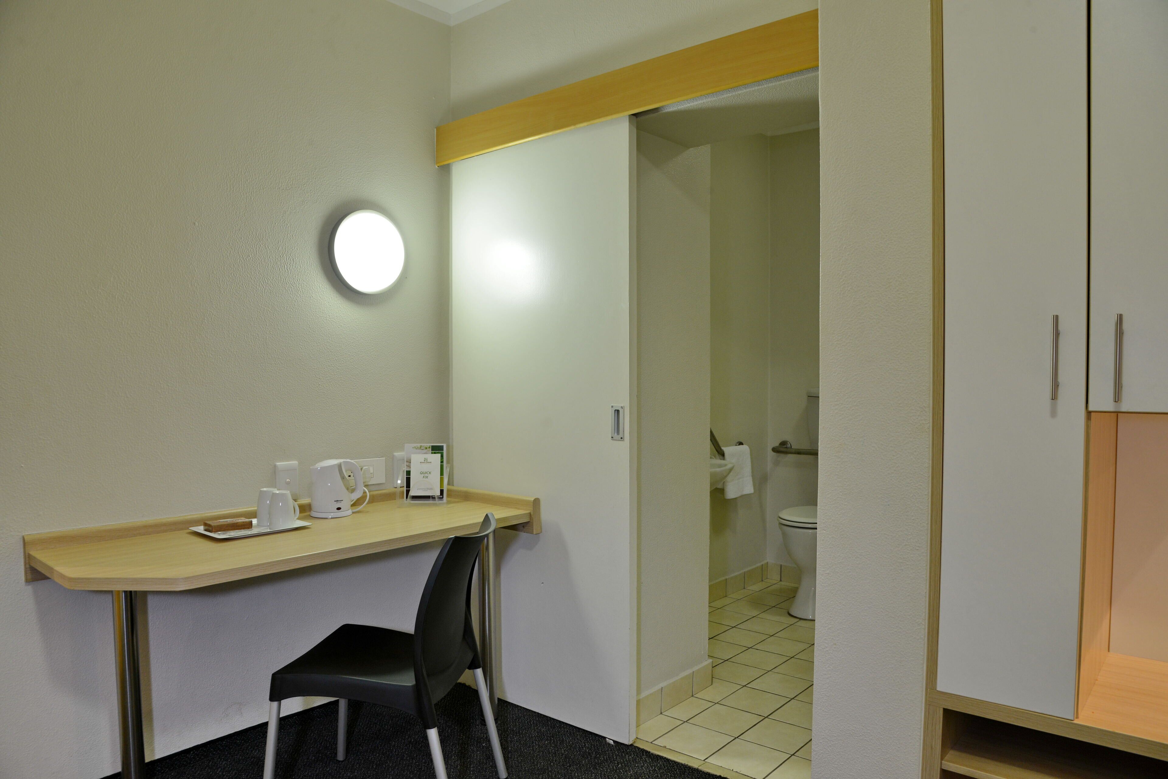 A small room with a desk, chair, and a round light fixture. There is a door leading to a bathroom with a toilet and sink.