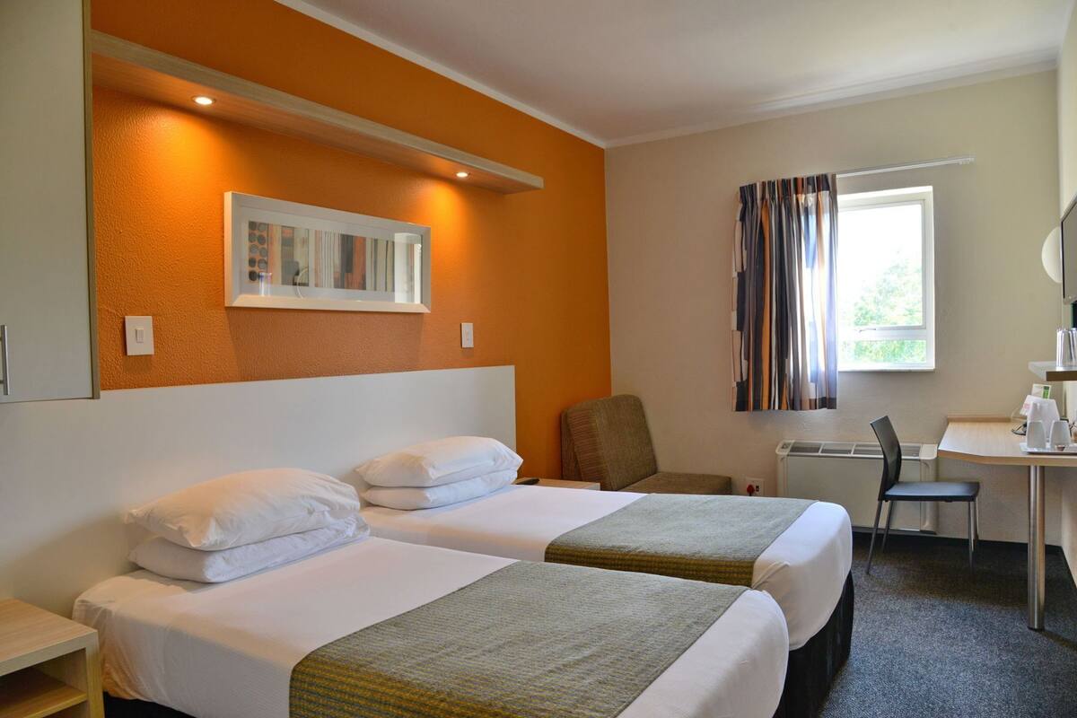A hotel room with two beds, a desk, a chair, a window with curtains, and a wall-mounted picture.