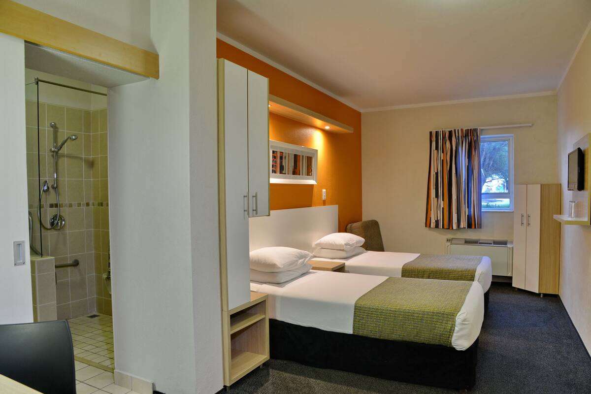 A hotel room with two beds, a shower, a small window with curtains, and a TV on the wall.