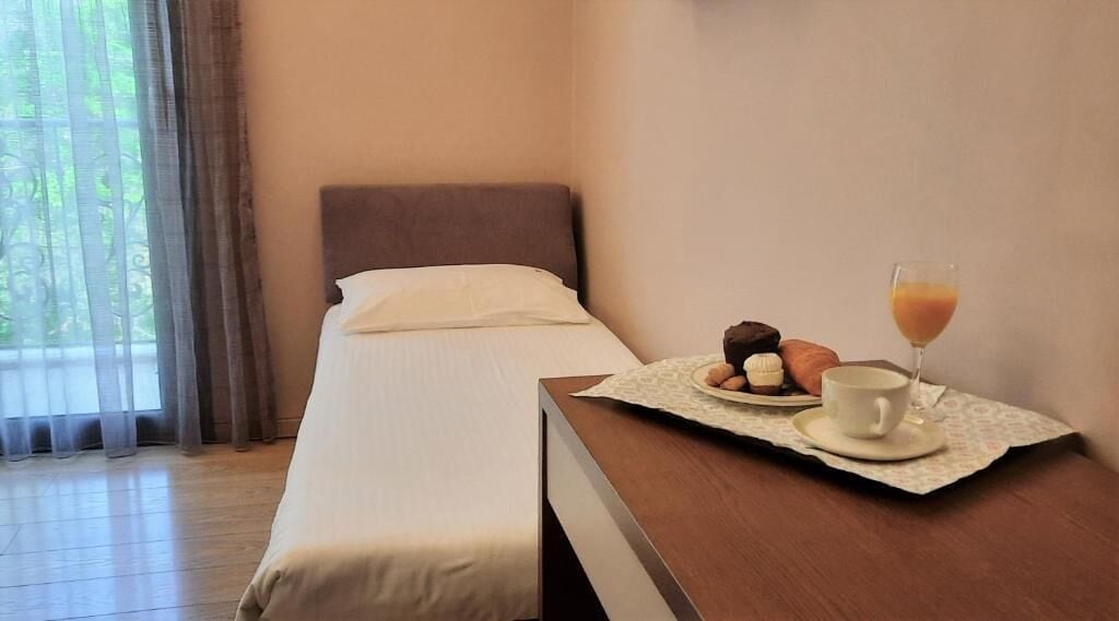A hotel room with a bed, a wooden nightstand, a breakfast tray with pastries and a glass of orange juice, and a window with curtains.