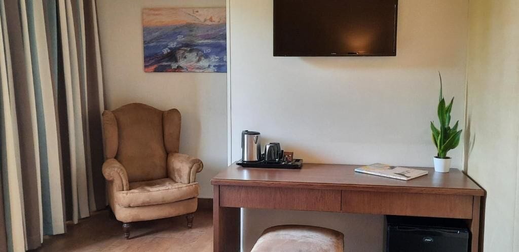 Una habitación con un escritorio de madera, un sillón color beige, una planta en maceta, un cuadro en la pared y un televisor montado sobre el escritorio.
