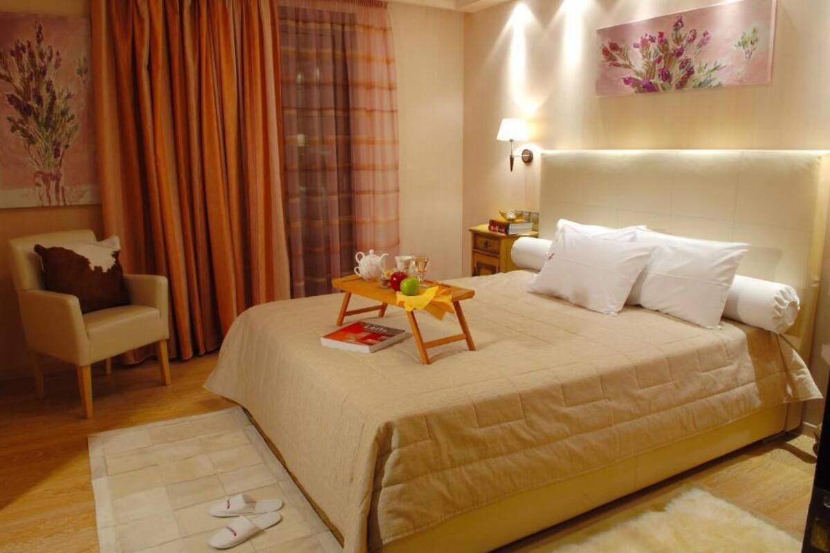 A hotel room with a large bed, a bedside table with a lamp, a chair, and a small table with a tray of food and a book.