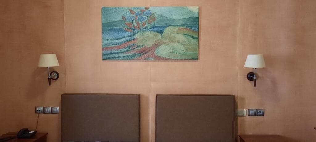 A hotel room with two beds, a painting on the wall, and two wall-mounted lamps.