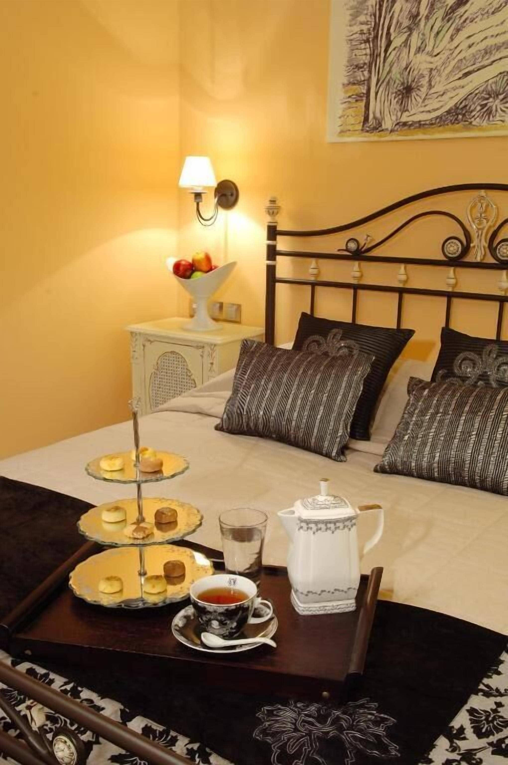 Una cama bien hecha con cabecera, una mesita de noche con lámpara, una bandeja con pasteles y una tetera, y una taza de té.