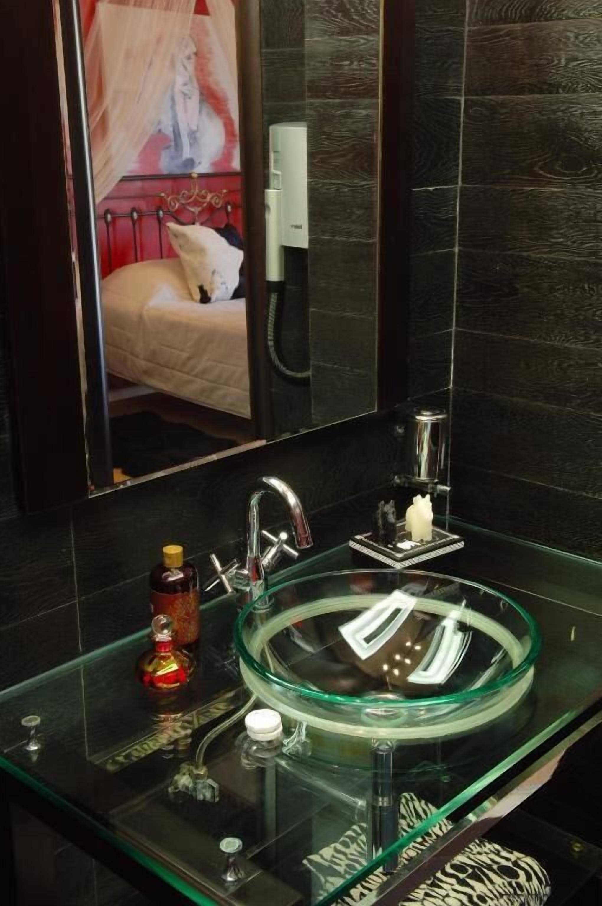 Un baño moderno con un lavabo de vidrio, un espejo y una cama que se ve en el reflejo.