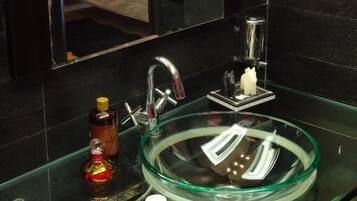 Un baño moderno con un lavabo de vidrio, un espejo y una cama que se ve en el reflejo.