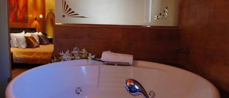 Una bañera ovalada grande con múltiples jets, un espejo encima y una cama con almohadones al fondo.
