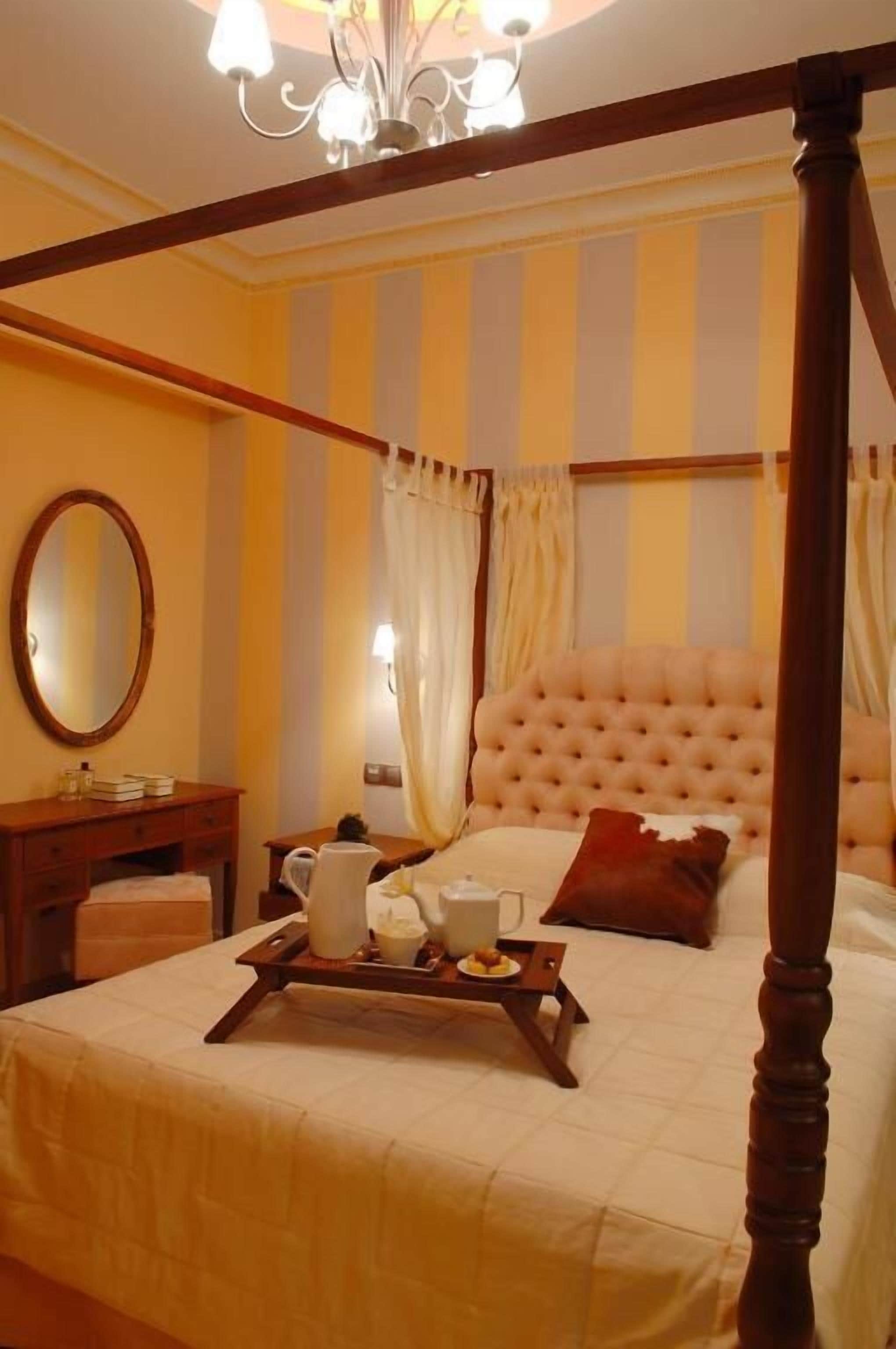 A four-poster bed with a tufted headboard, a bedside table with a mirror, and a tray with breakfast items.