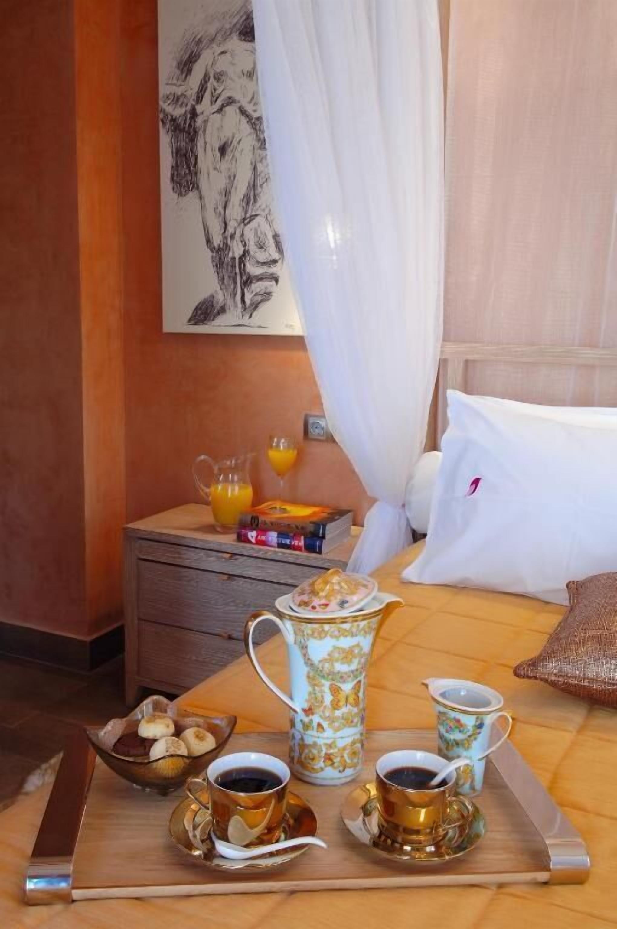 A neatly arranged breakfast tray with coffee and tea on a wooden table, a bowl of fruit, and a pitcher of orange juice on a bedside table.