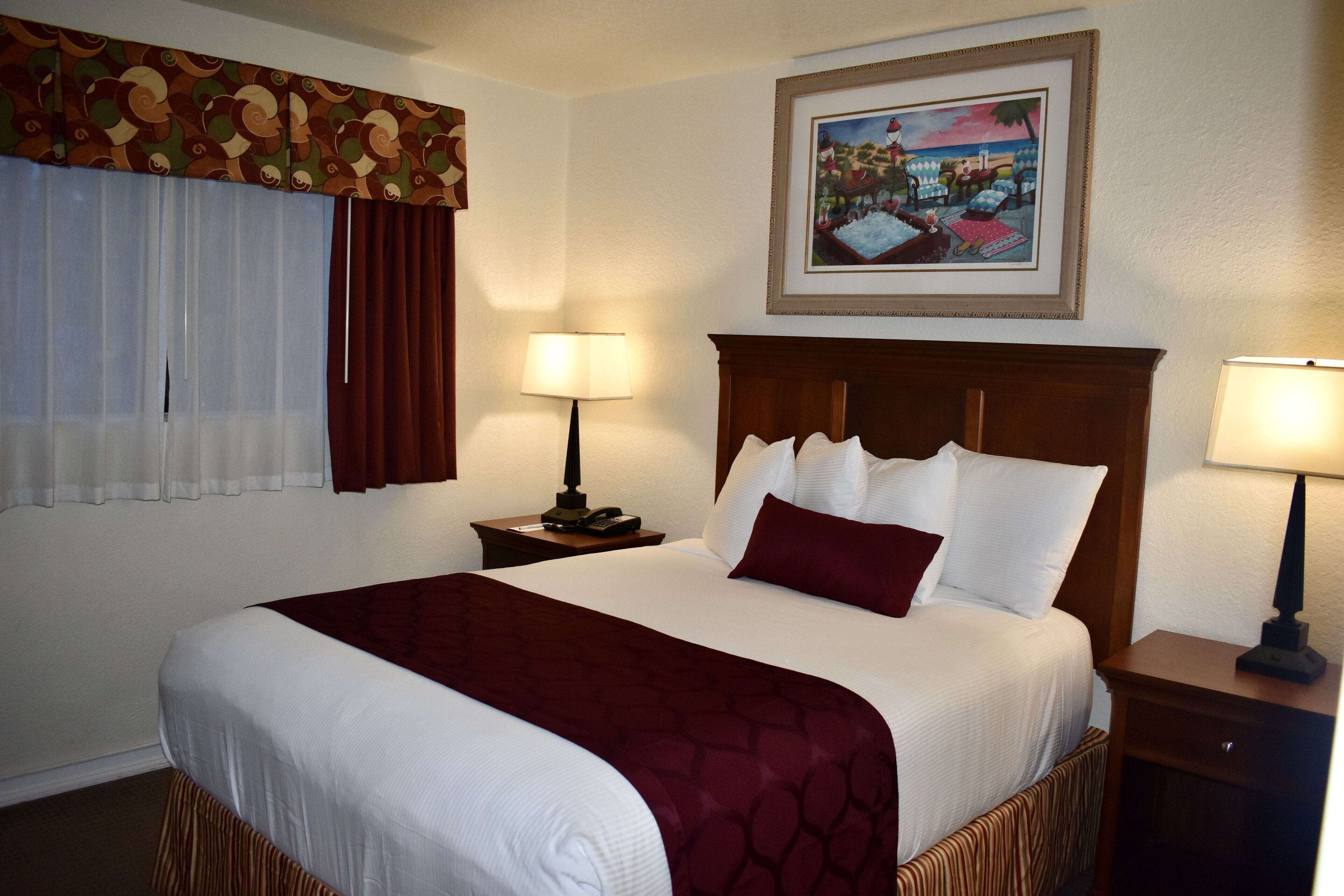 A hotel room with a large bed, a nightstand with a lamp, a framed picture on the wall, and a window with curtains.