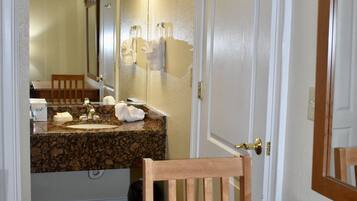 Un baño con lavamanos, espejo y tres lámparas encima. Hay una silla de madera y una mesa en la habitación.