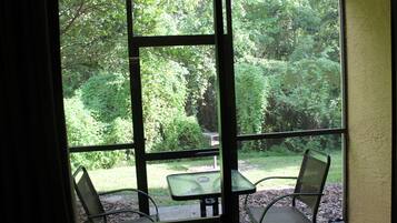 Quarto com uma grande janela que oferece vista para um jardim exuberante. Há uma pequena mesa e duas cadeiras no interior.
