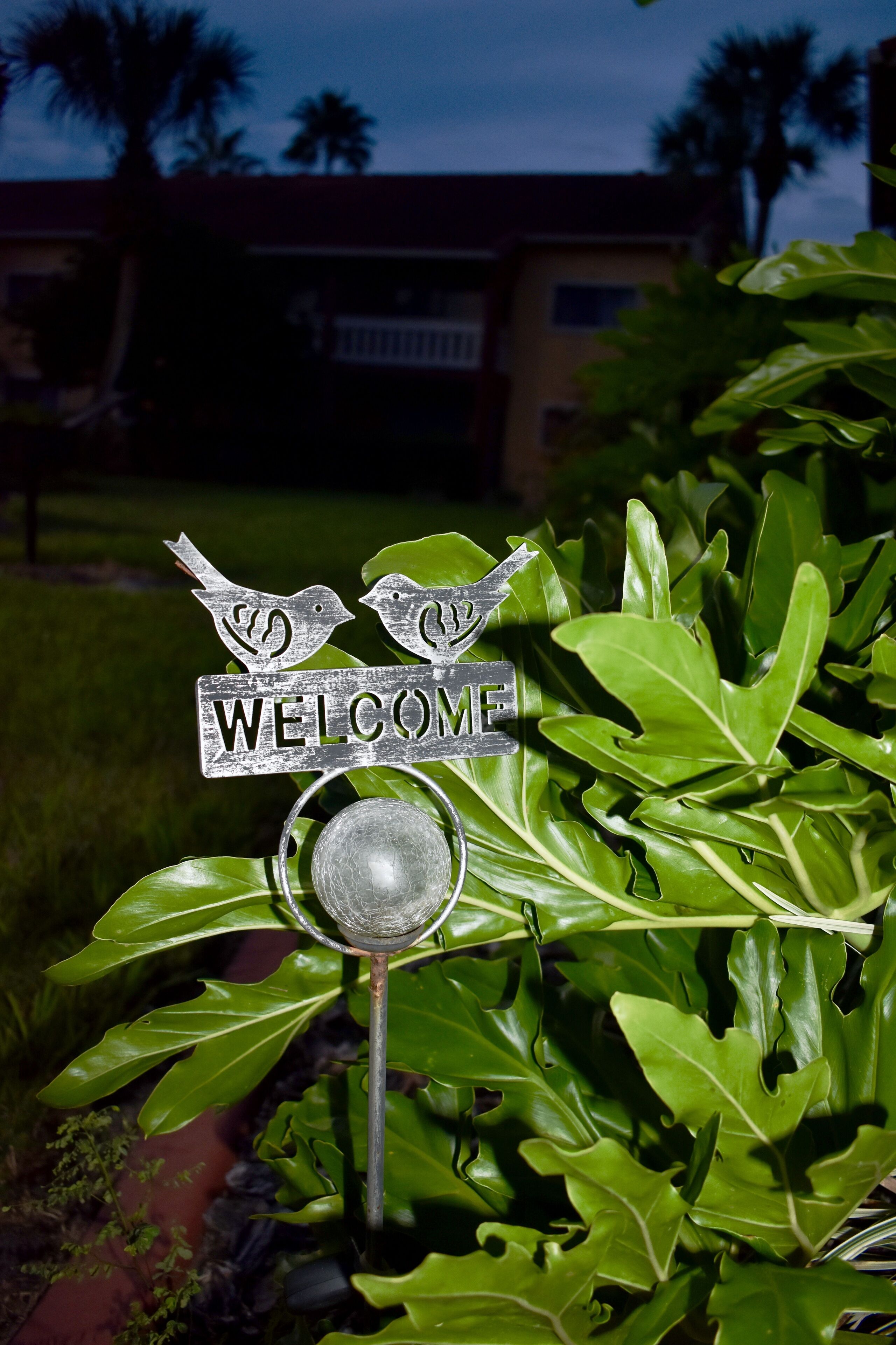 Señal de bienvenida con siluetas de aves y la palabra 'WELCOME' en un jardín con follaje verde.