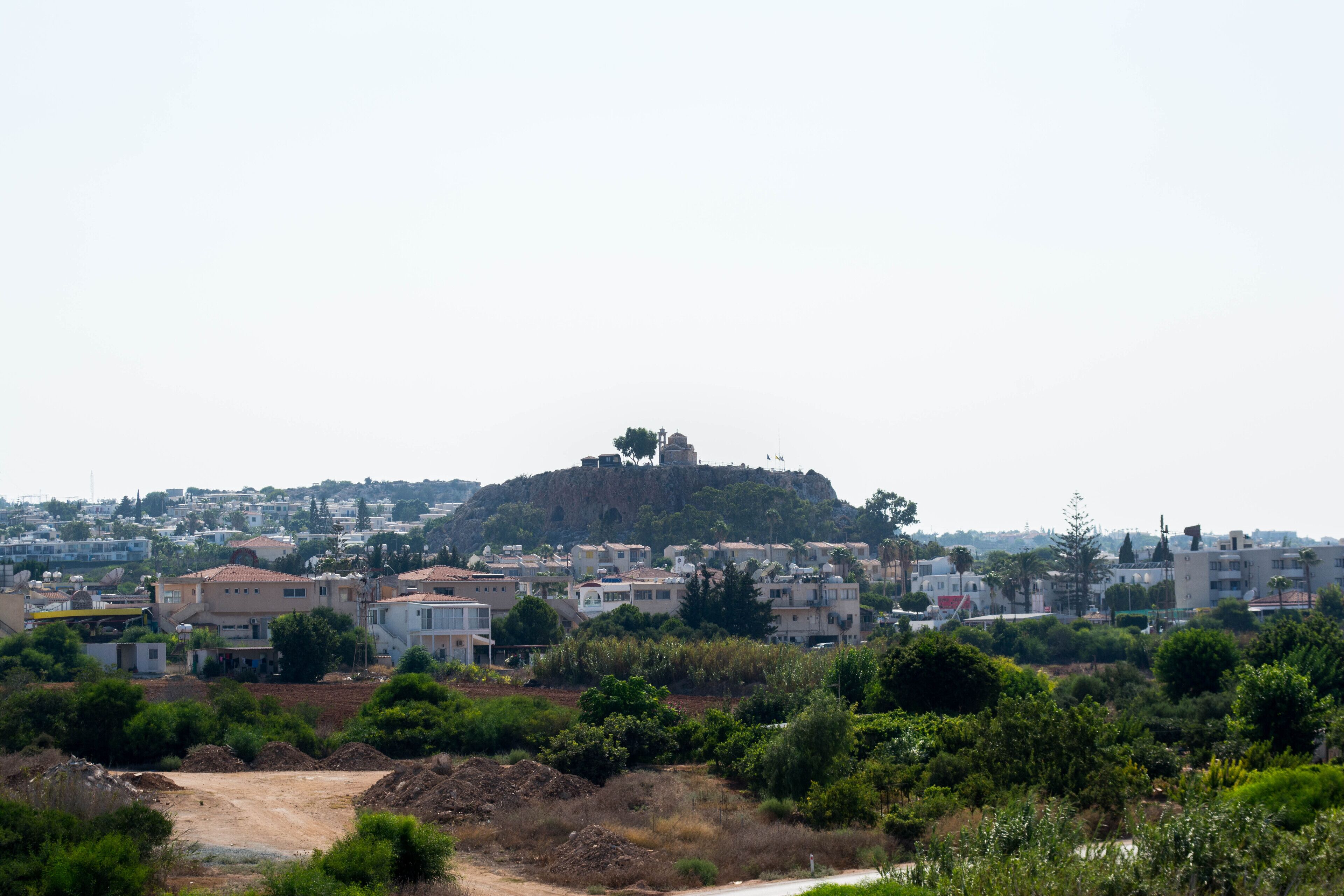 A residential area with houses on a hill, a dirt road, and greenery.