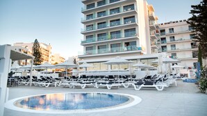 Un espace aménagé au bord de la piscine, avec des chaises longues et des parasols, un bâtiment moderne doté de balcons, et un ciel dégagé.