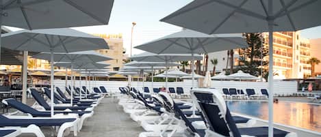 A row of sun loungers with umbrellas, a pool, and a building in the background.