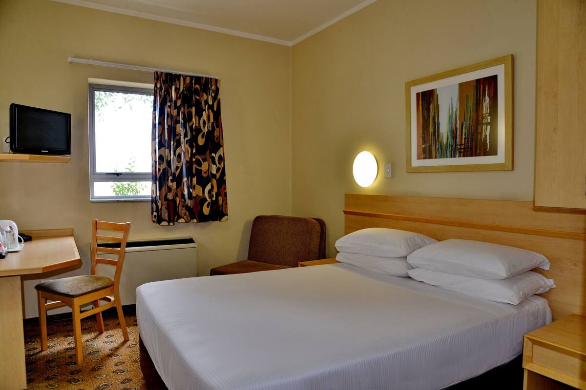 Een hotelkamer met een bed, een bureau met stoel, een televisie, een raam met gordijnen en een schilderij aan de muur.