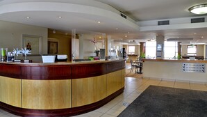 En hotellobby med en böjd receptionsdisk i trä, en sittgrupp med stolar och en skylt som visar rumens nummer och telefonnummer.