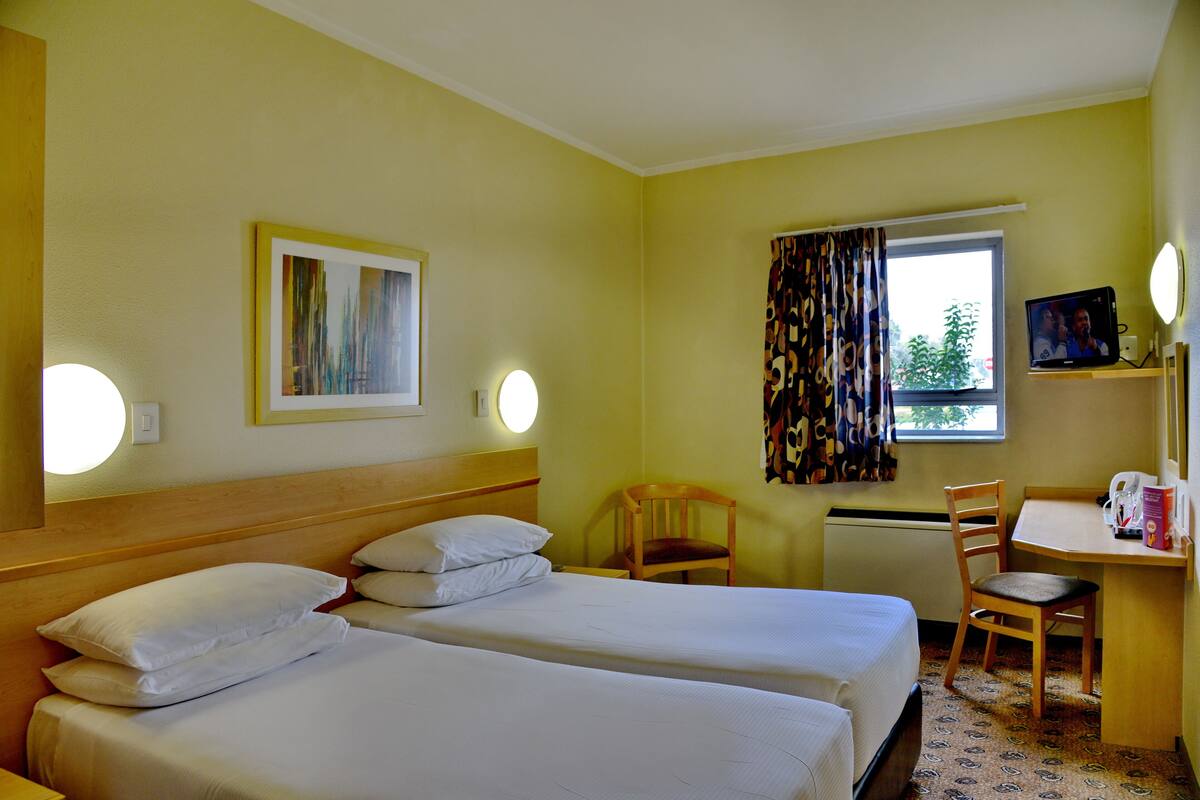 Een hotelkamer met twee bedden, een bureau, een stoel, een raam met gordijnen, een schilderij aan de muur en een televisie.