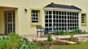 Un edificio con ventanal saliente, una puerta y un pequeño patio con sillas y plantas.