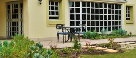A building with a bay window, a door, and a small patio area with chairs and plants.