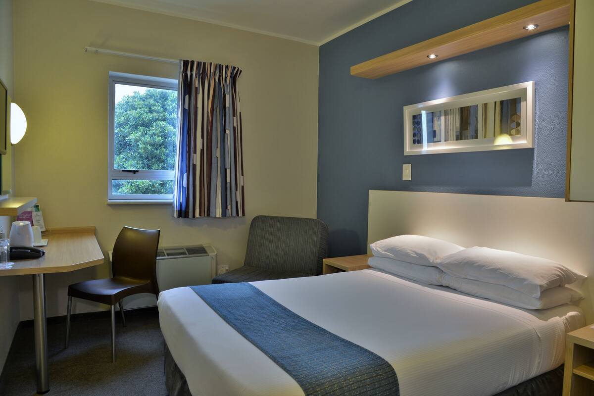 Una camera d'albergo con un letto, una scrivania, una sedia, una finestra con tende e uno specchio a parete.