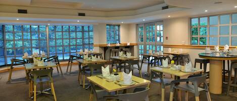 A restaurant with multiple tables and chairs, large windows with blinds, and a carpeted floor.