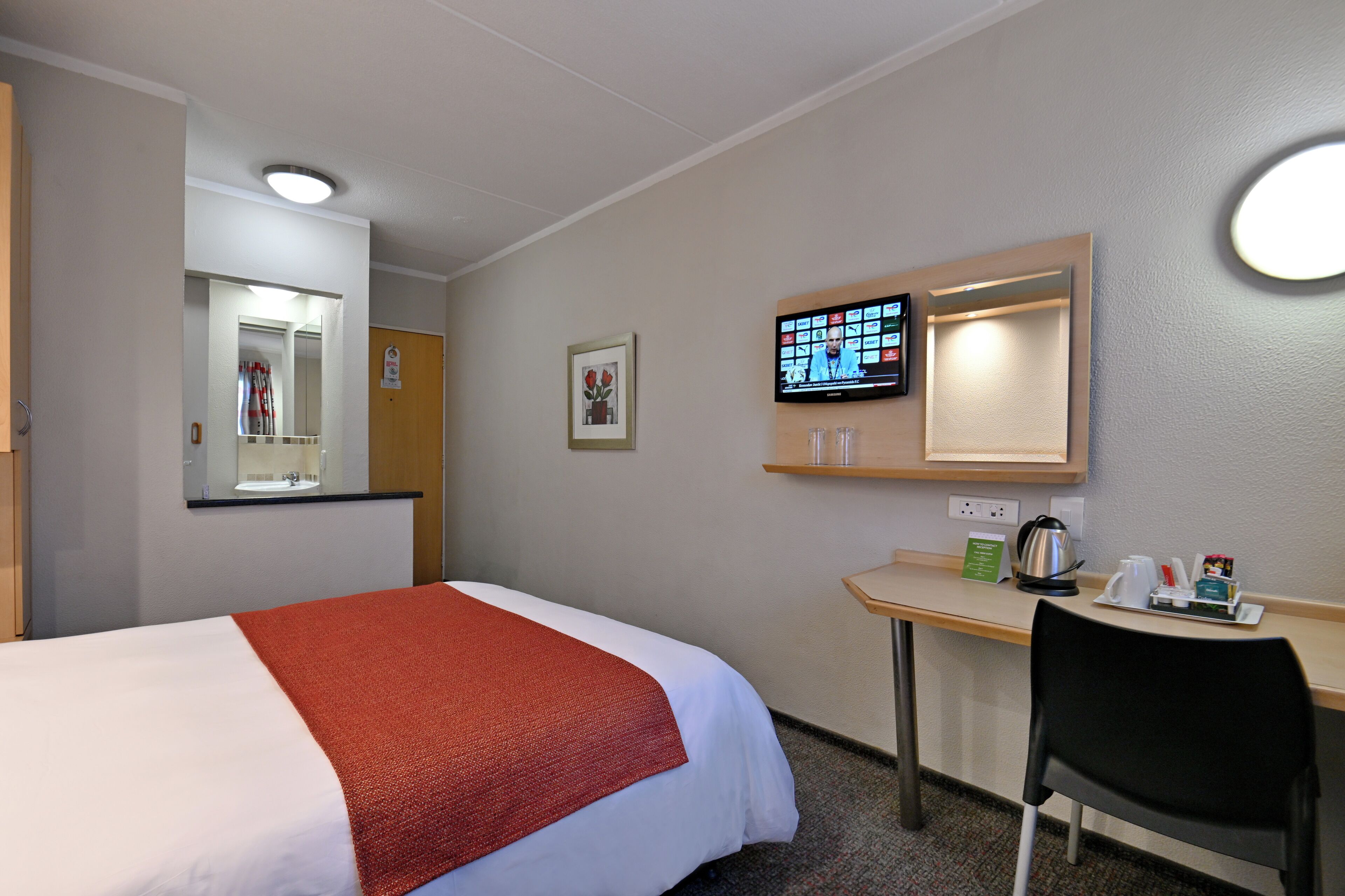 Una habitación de hotel con cama, escritorio, silla, televisor y espejo.