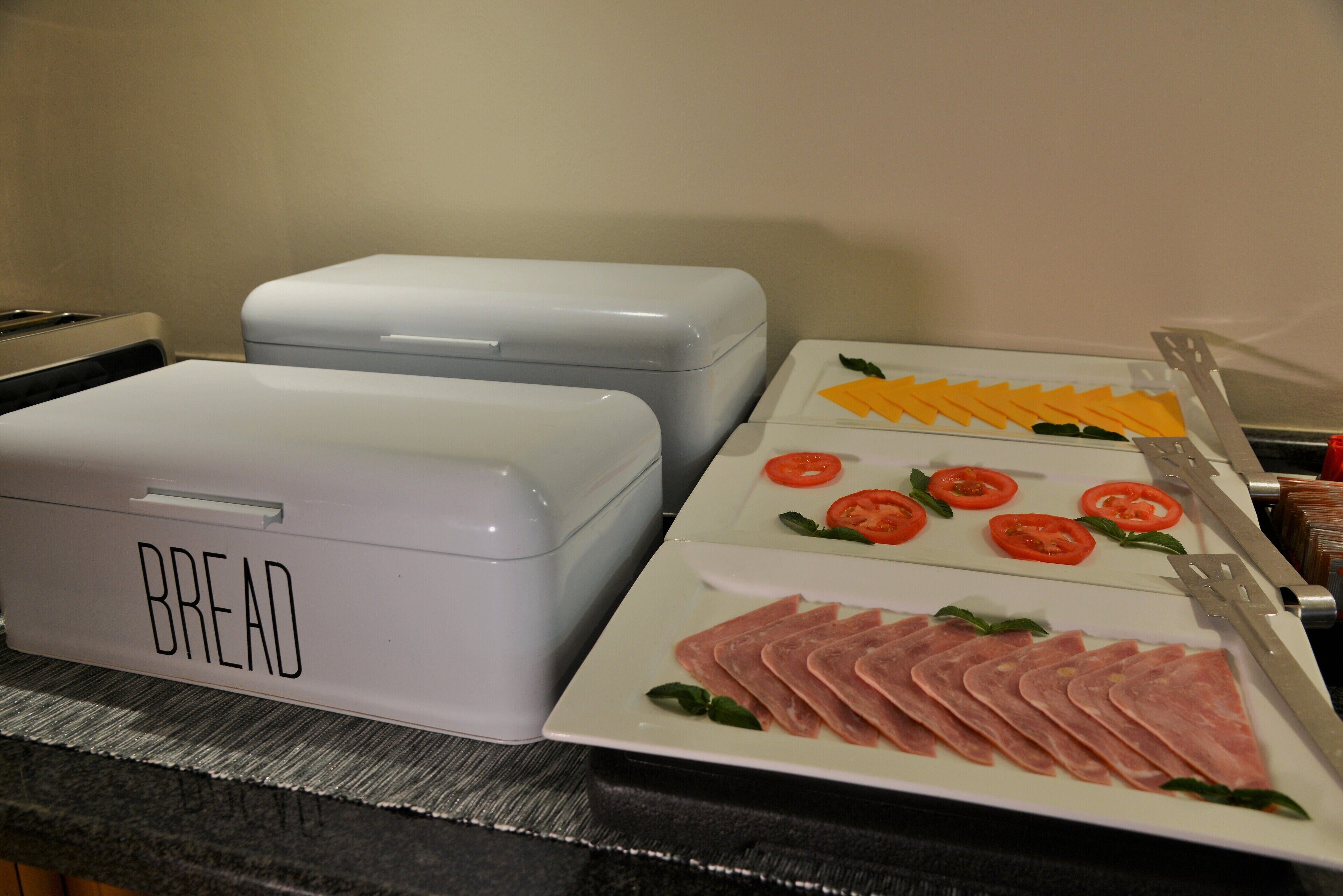 Una encimera de cocina con recipientes etiquetados para pan y un plato con jamón y tomates en rodajas.