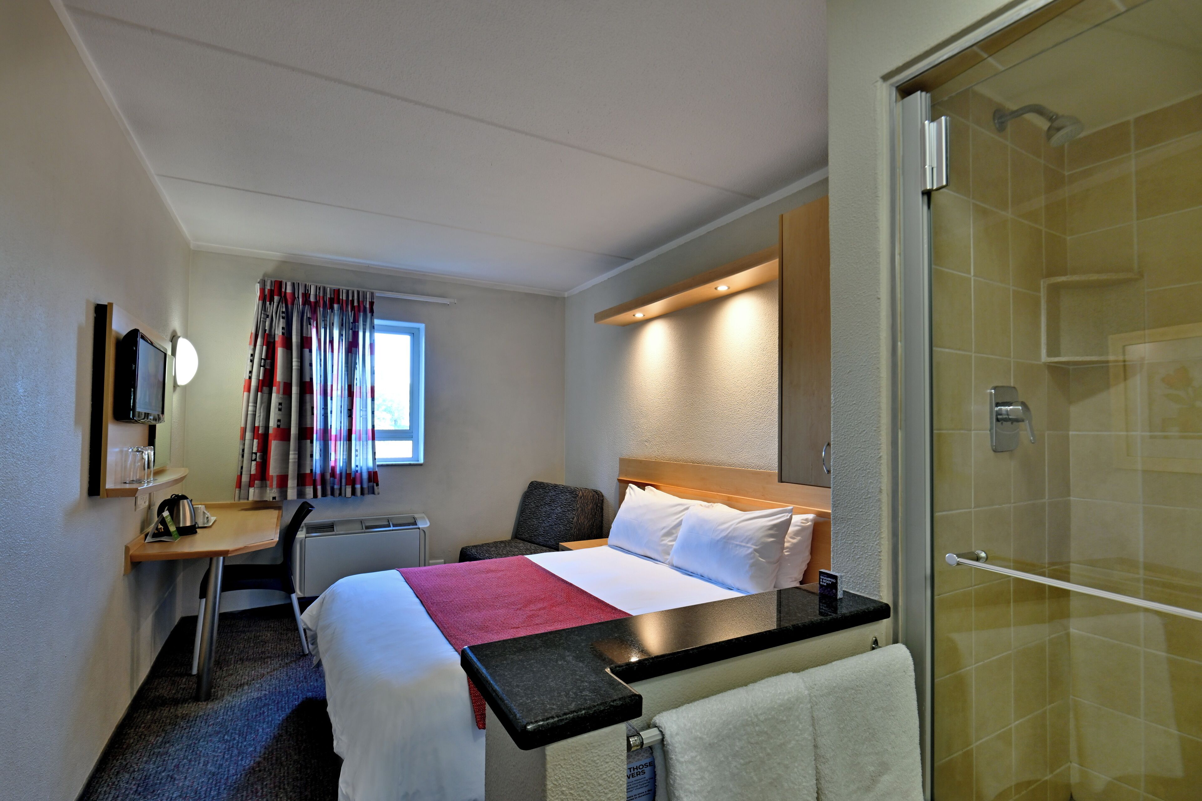 Una habitación de hotel con cama, mesita de noche, escritorio, silla y zona de ducha.