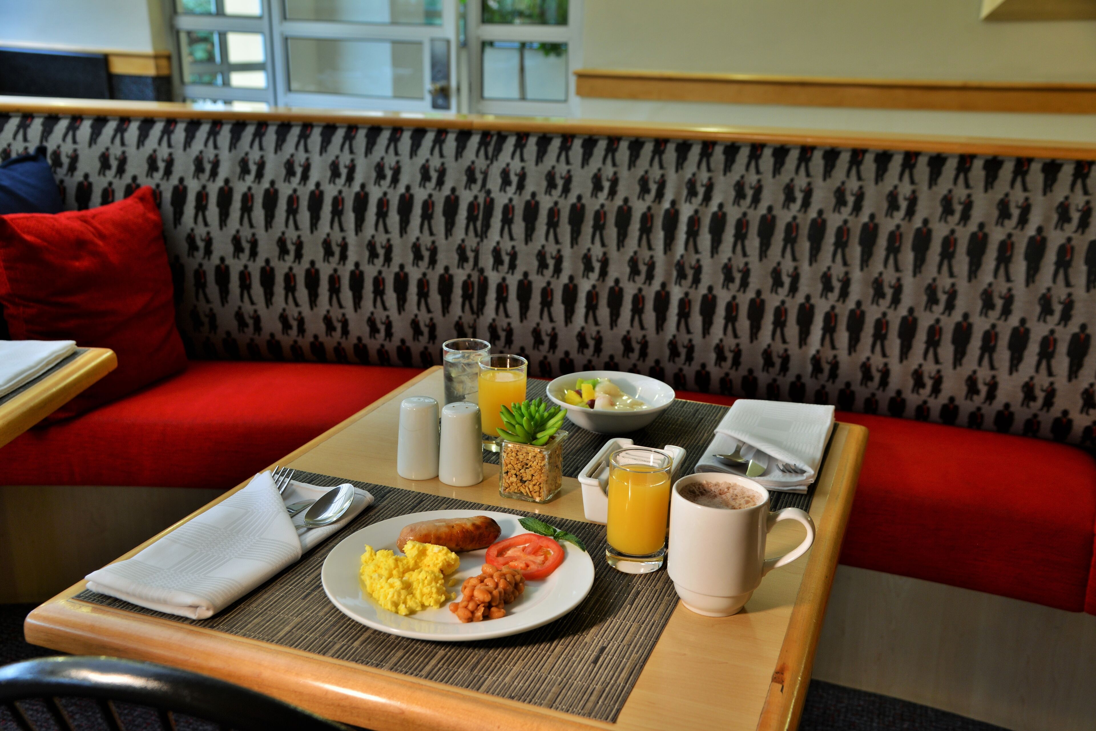 Une table en bois est dressée pour le petit-déjeuner, avec une assiette d’œufs brouillés, des haricots, des tomates et des saucisses, un verre de jus d’orange, une tasse de café et un bol de fruits.