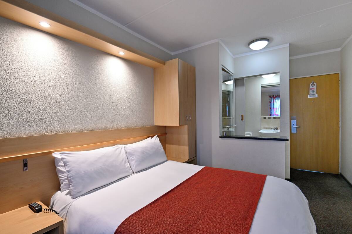 A hotel room with a bed, a nightstand, a wooden headboard, a red throw blanket, a mirror, a sink, and a door.