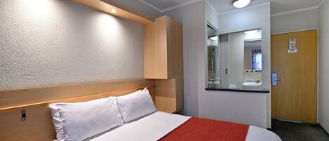 Habitación de hotel con cama, mesita de noche, cabecero de madera, manta roja, espejo, lavamanos y puerta.