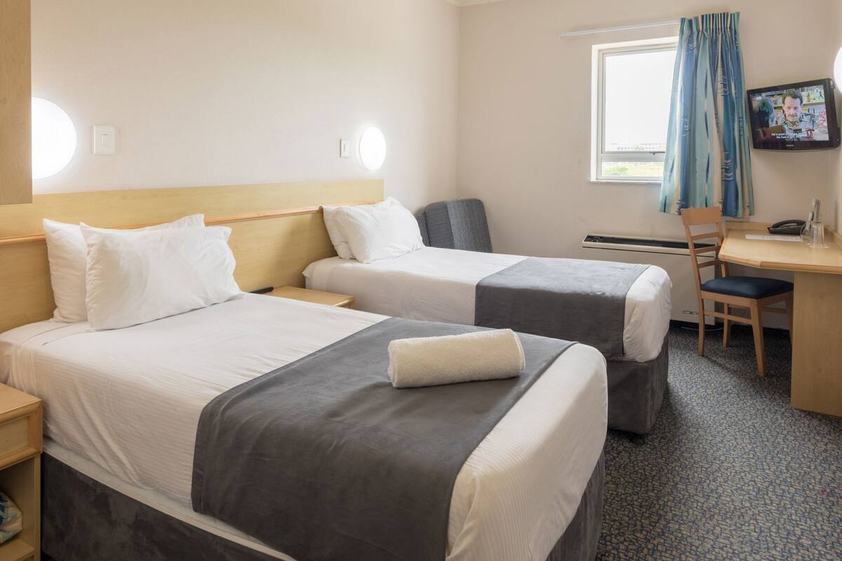 Une chambre d’hôtel avec deux lits, un bureau, une chaise, une télévision et une fenêtre avec des rideaux.