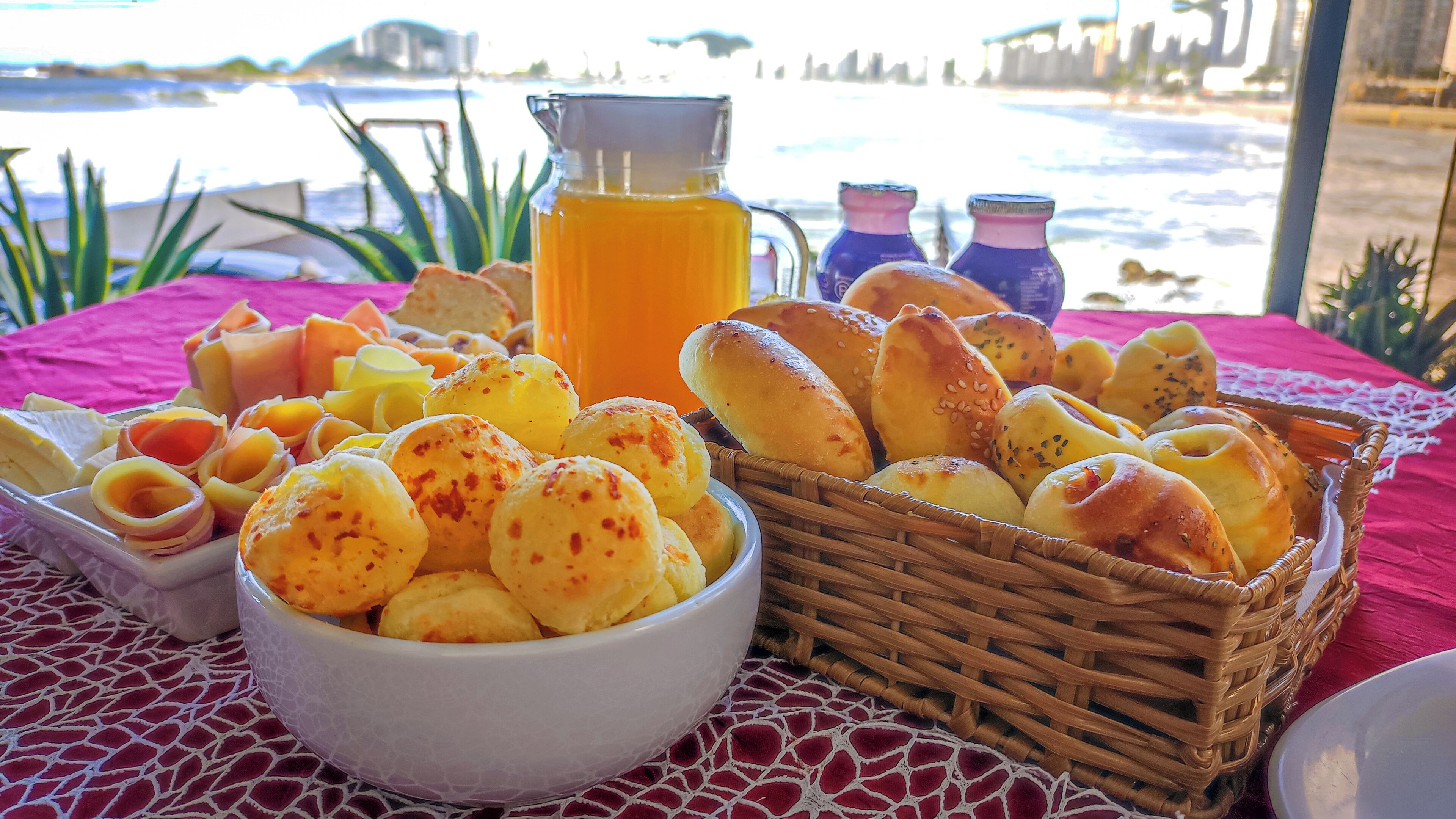 Una mesa con diversos alimentos, una jarra de jugo de naranja y botellas de bebidas, frente a un telón de fondo con vista a la playa y al océano.