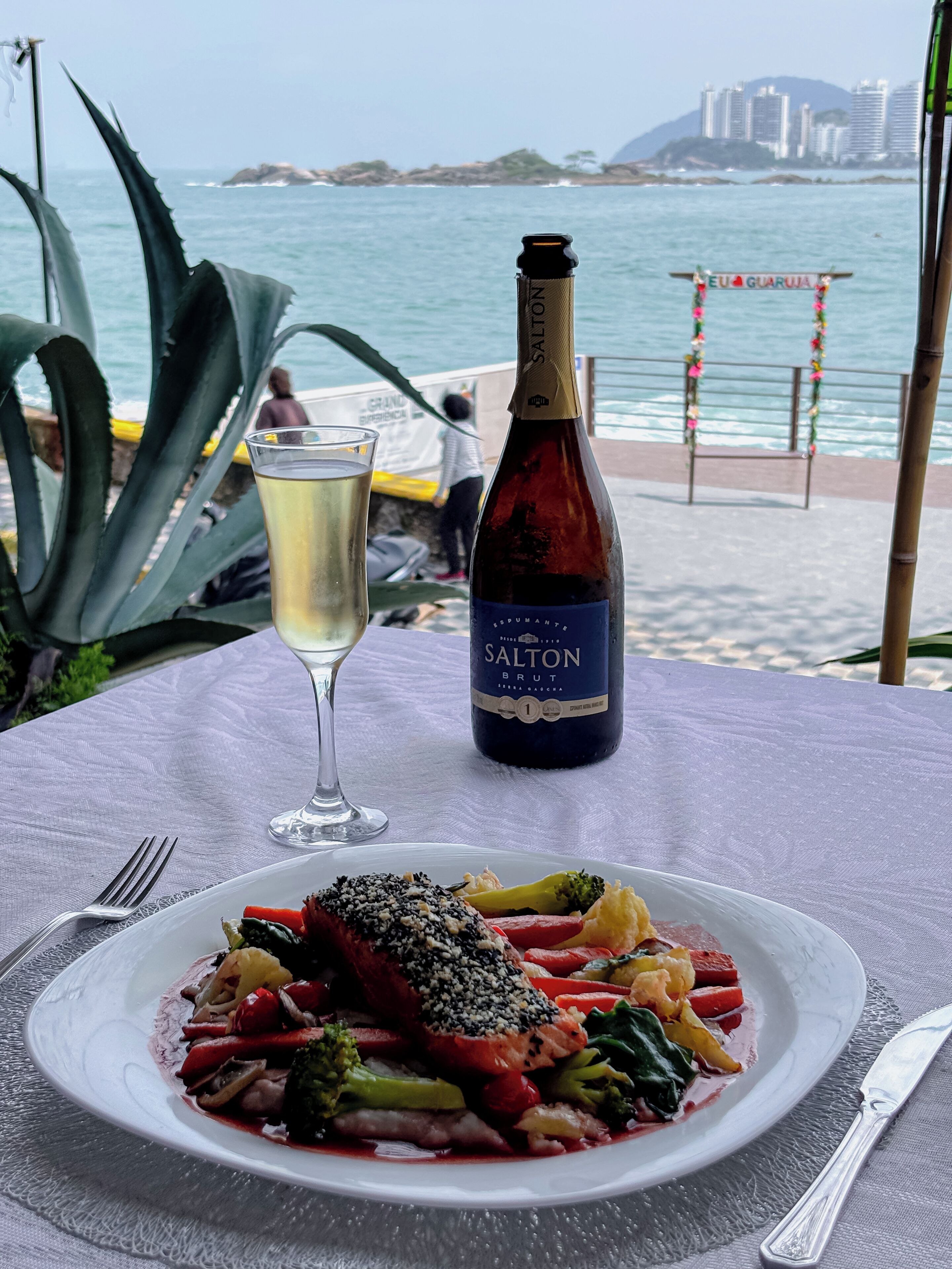Una mesa con un plato de comida, una botella de vino y una copa de vino blanco, con vista al mar y edificios al fondo.