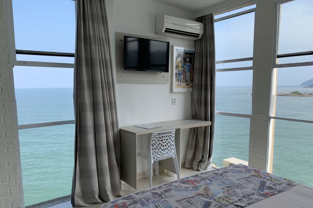 A room with a bed, a desk, a TV, and a large window overlooking the sea.