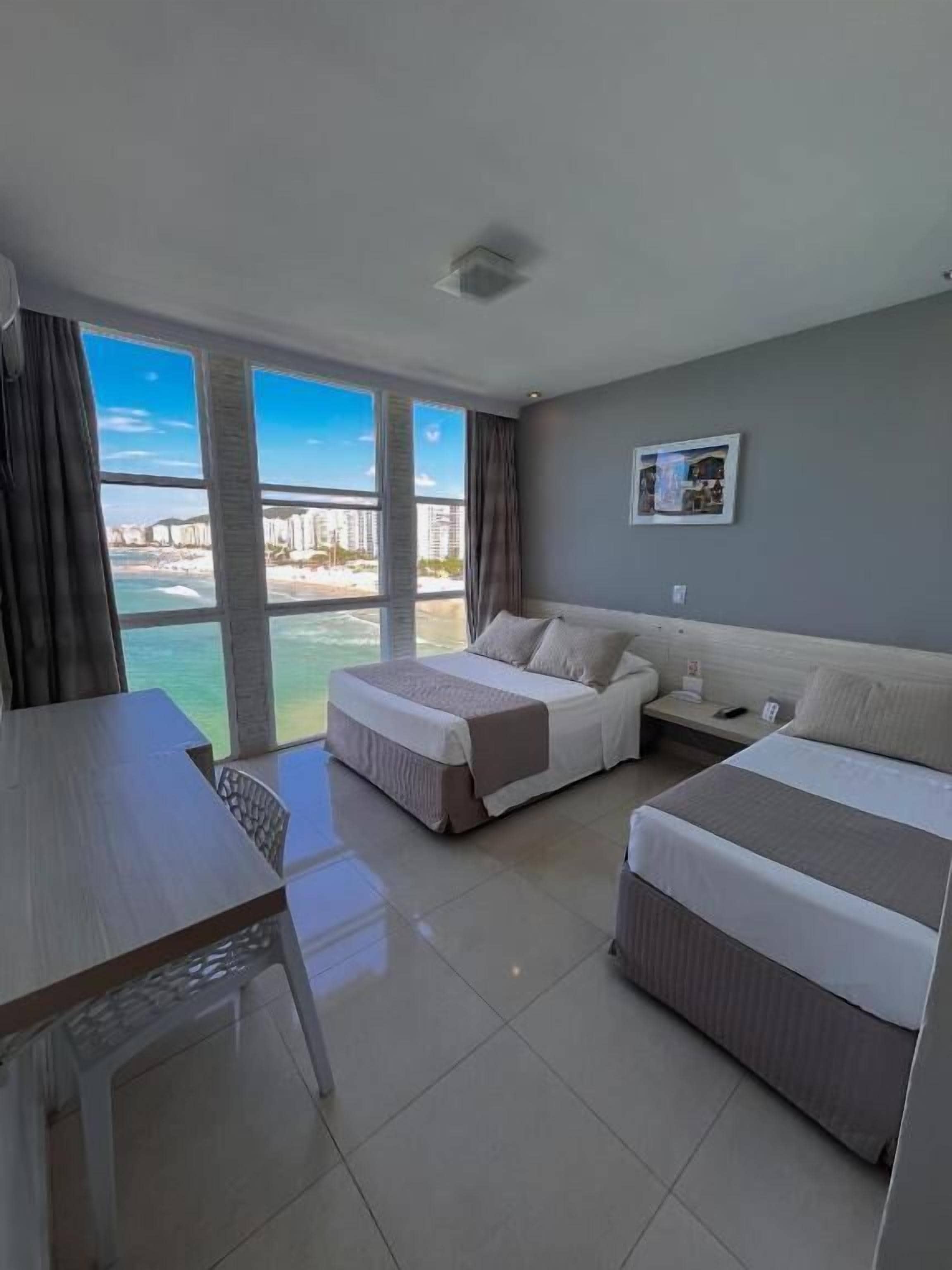 A hotel room with two beds, a desk, and a view of the sea.