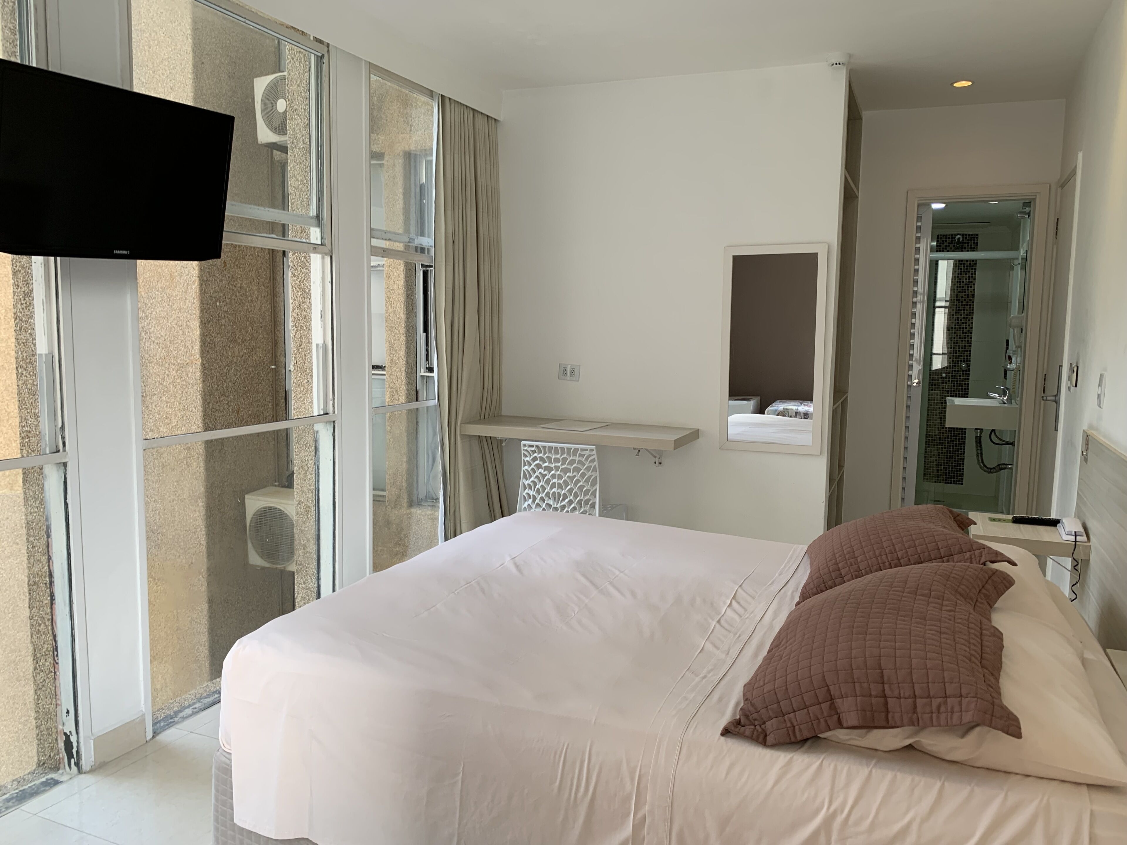 Quarto de hotel com uma cama grande, televisão de tela plana, banheiro com porta de vidro e uma pequena escrivaninha com espelho.