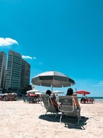 Una playa con tumbonas y sombrillas, un cielo azul despejado y edificios altos al fondo.