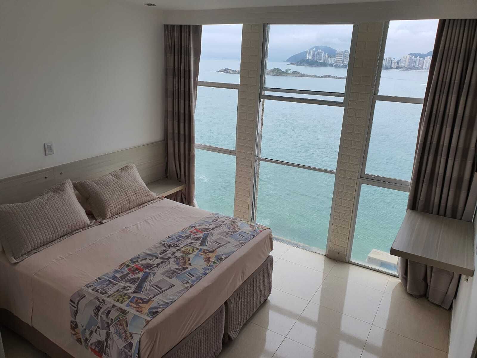 A bedroom with a large window, a bed with a patterned bedspread, and a view of a body of water and buildings.