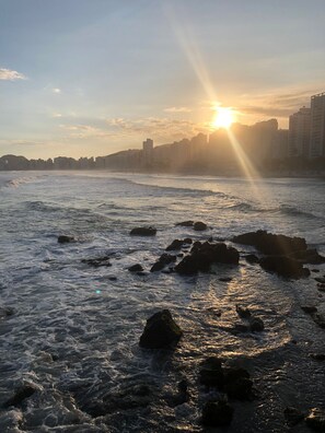 Uma praia com ondas e rochas, silhueta urbana ao fundo, e o sol se pondo ou nascendo.