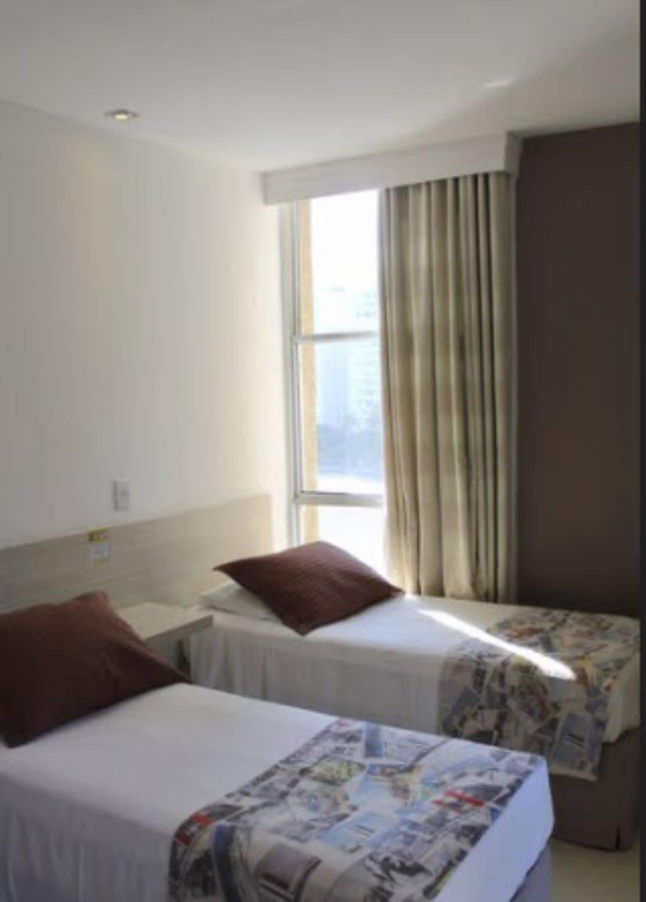 Ein Hotelzimmer mit zwei Einzelbetten, einem fenster mit Vorhängen und Blick auf die Stadtlandschaft.