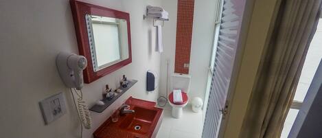 A bathroom with a red sink, a mirror, a hairdryer, a toilet, and a window with curtains.