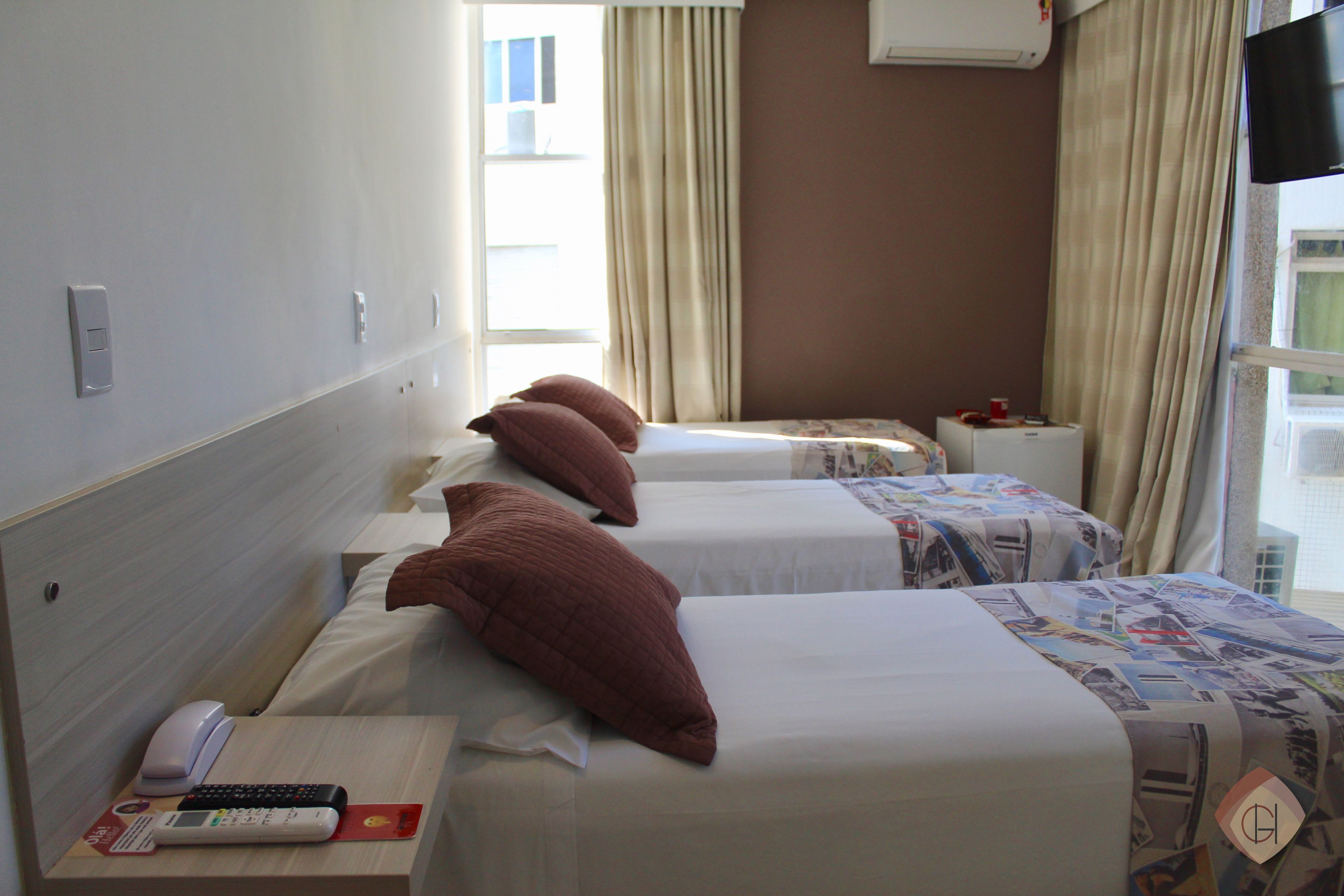 A hotel room with three single beds, each with a pillow and a blanket. There is a bedside table with a phone and a remote control.