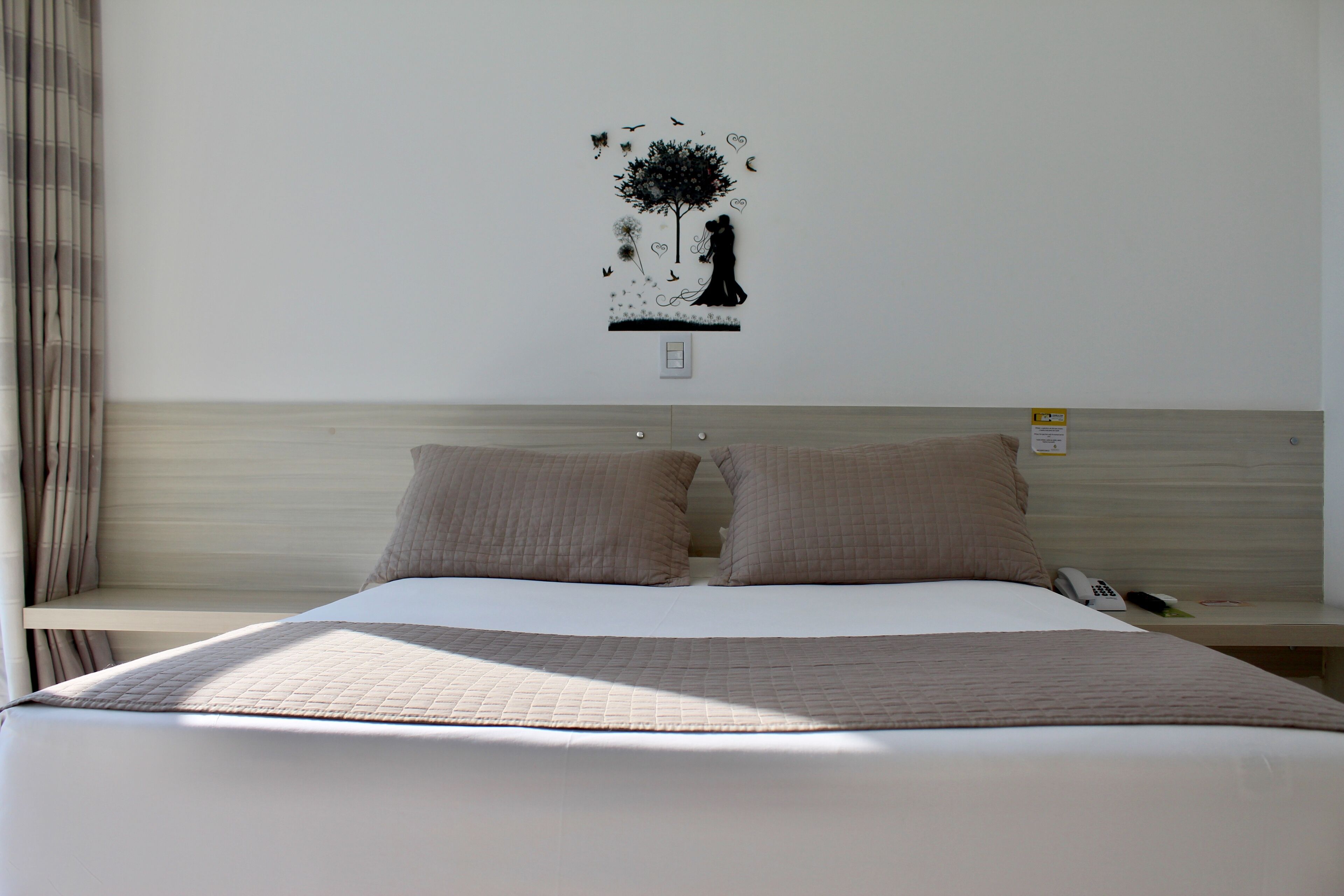 Une chambre d’hôtel avec un lit, deux oreillers, une table de chevet avec un téléphone et une décoration murale représentant un arbre et un couple.