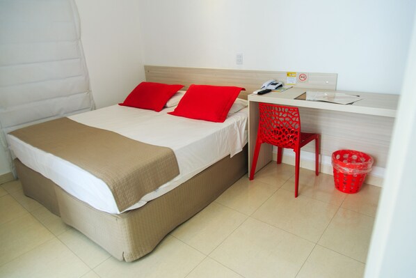 Una cama con cubierta color beige, dos almohadas rojas, un cabecero de madera, un escritorio con una silla roja y un bote de basura rojo.