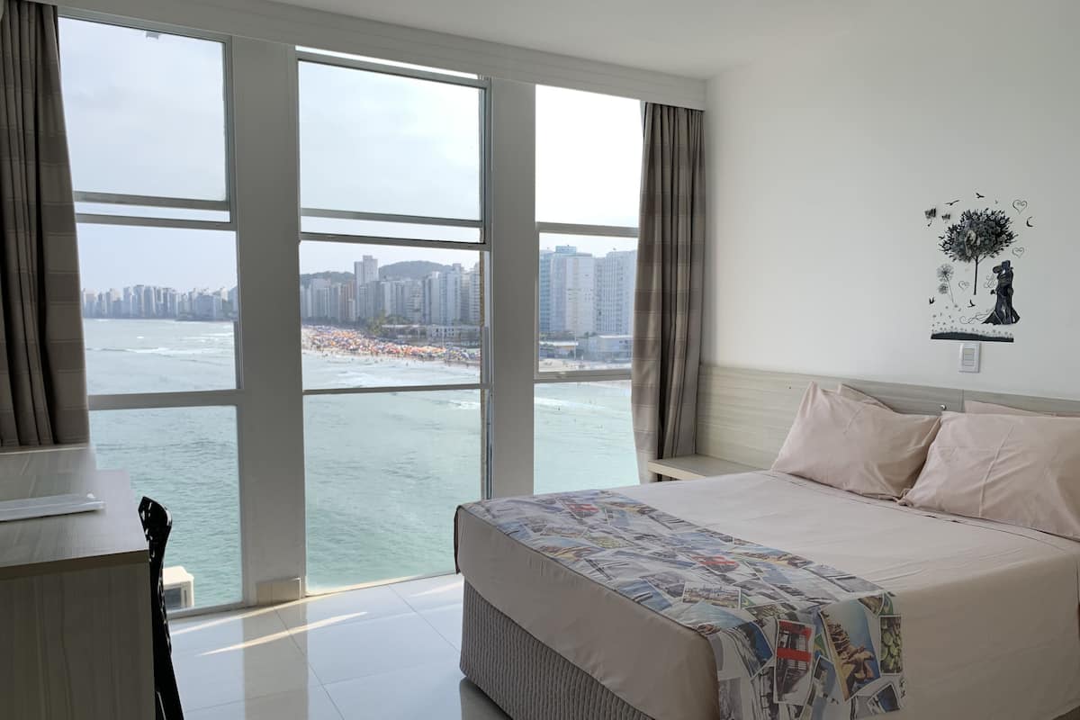 A hotel room with a large bed, a desk, and a view of a beach and cityscape through large windows.