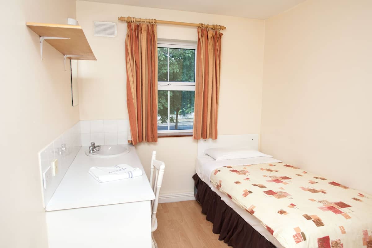 Quarto pequeno e limpo, com uma cama, mesa de cabeceira, espelho e janela com cortinas.