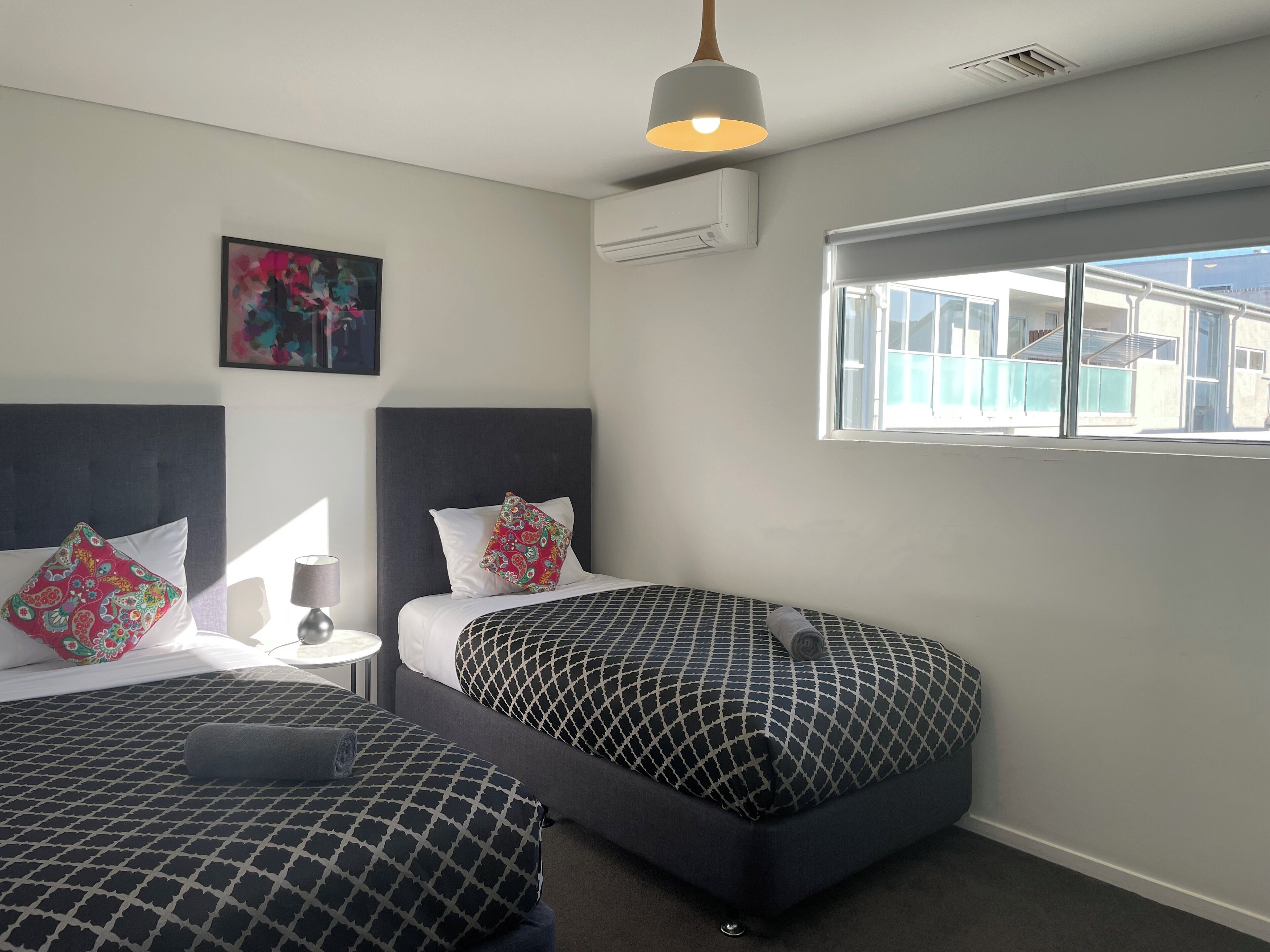 A modern hotel room with two beds, a large window, and a ceiling light.